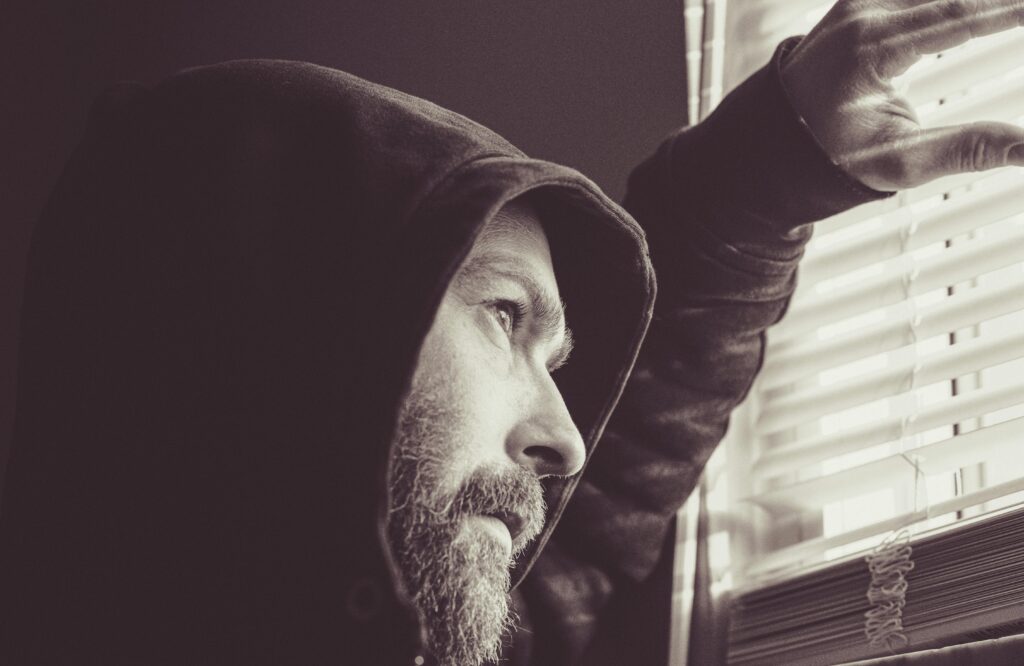 A bearded man in a hoodie looks thoughtfully out a window with blinds.