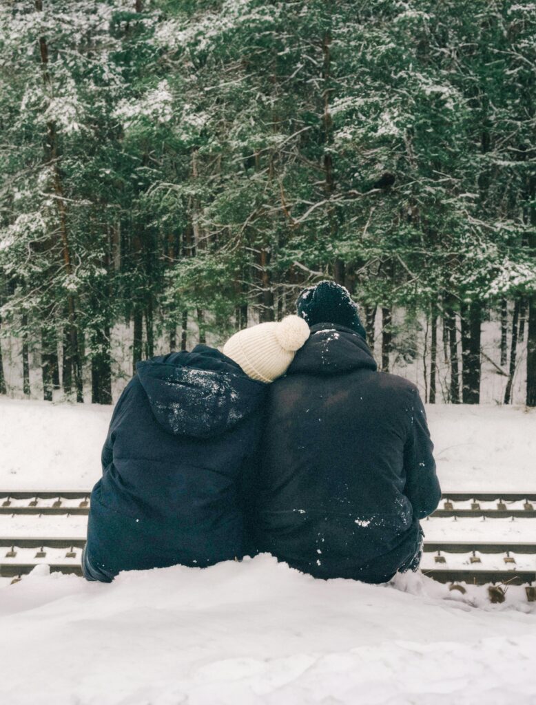 Couple in winter coats enjoy a snowy forest view sitting on railroad tracks.