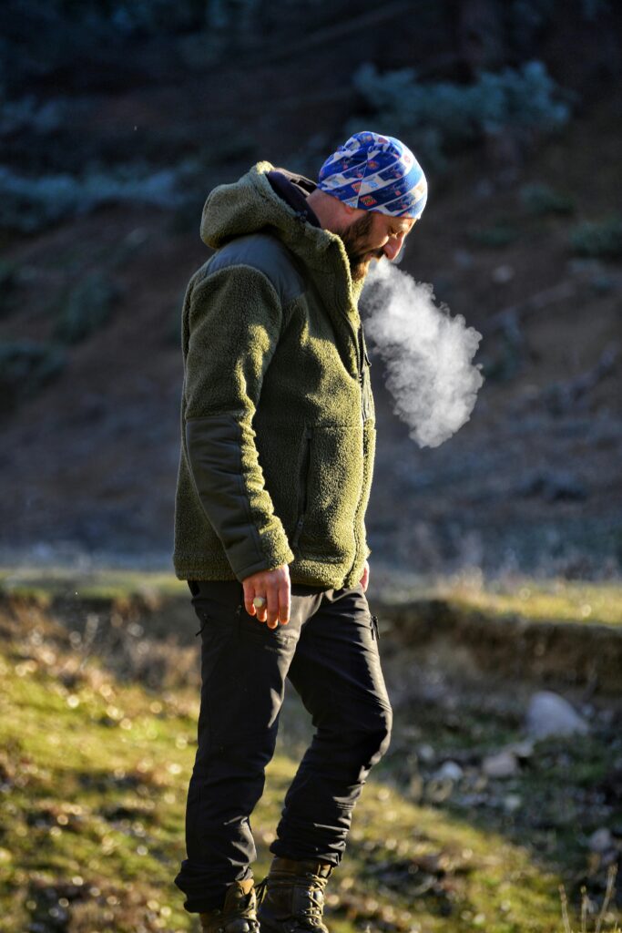 A man in a green jacket exhales misty breath in a chilly outdoor setting, highlighting a winter atmosphere.