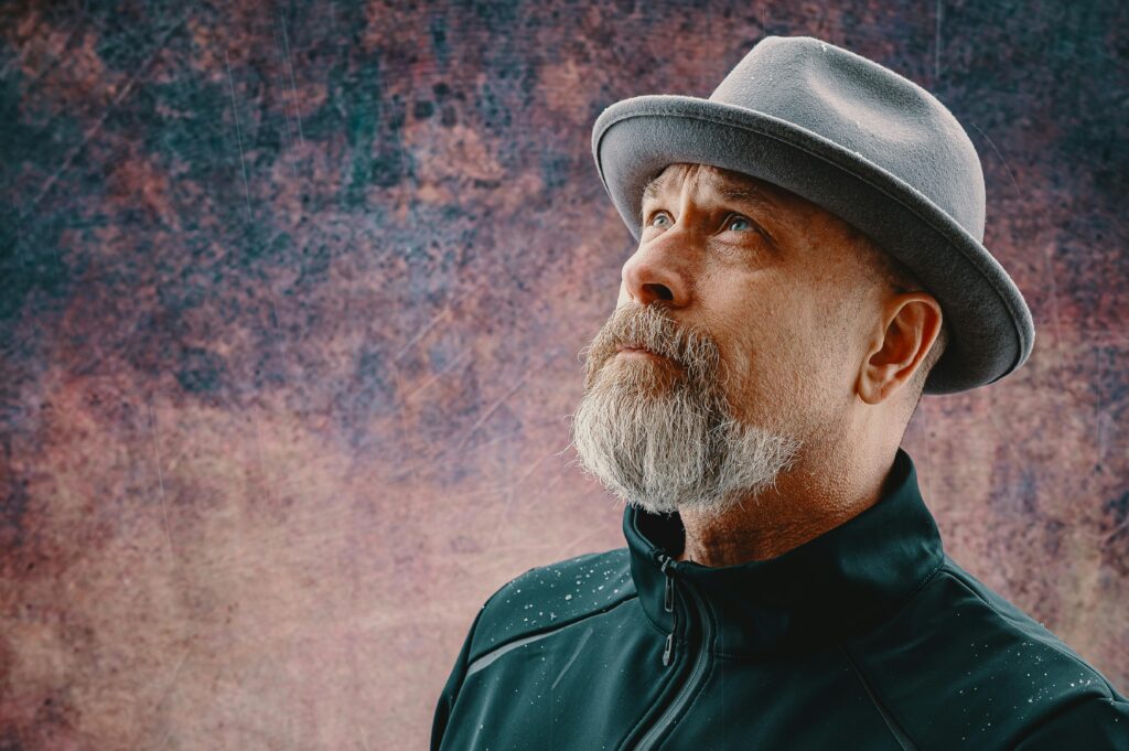 Close-up portrait of an elderly man with a beard wearing a fedora against a textured background.
