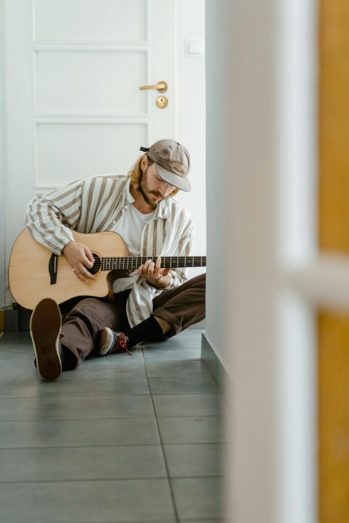 A man sits on the floor playing acoustic guitar in a bright room, enjoying leisure time.