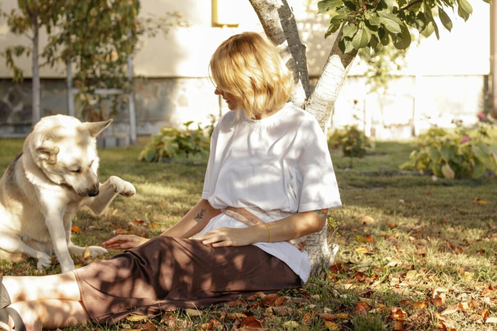 A woman enjoys a sunny day sitting under a tree with her dog in a serene garden.