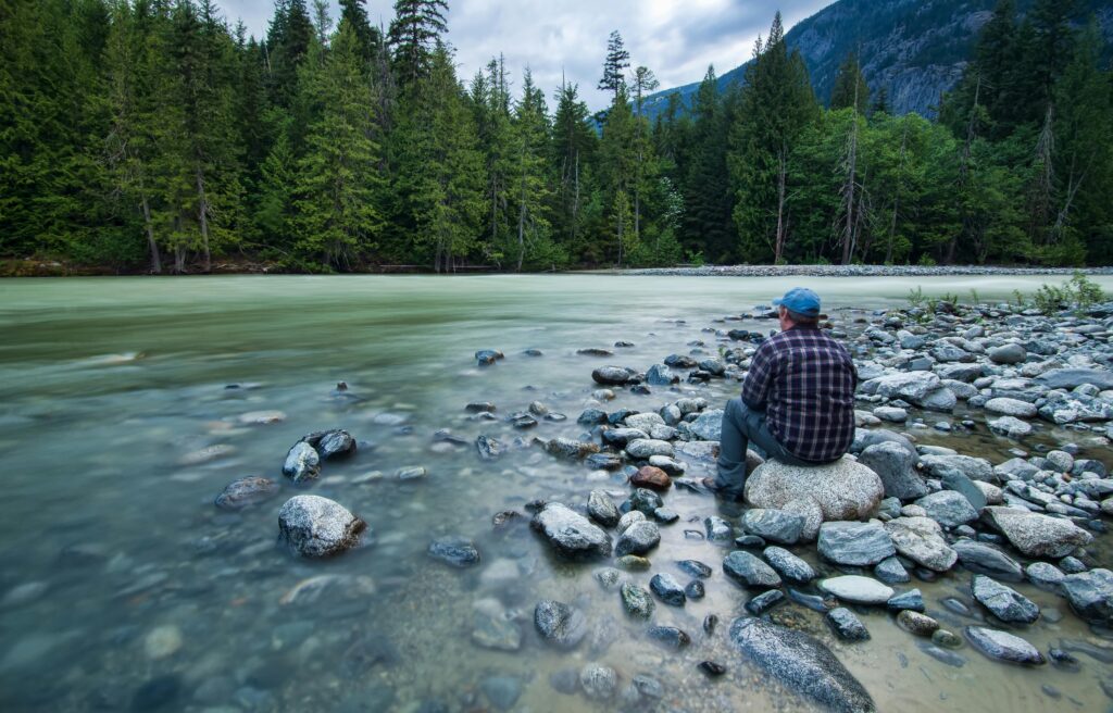 Man sitting by river in a forest setting in Pemberton, BC, Canada, surrounded by mountains.