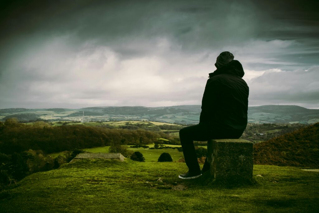A person in a jacket sits on a stone, gazing over a lush, rolling landscape under a cloudy sky.