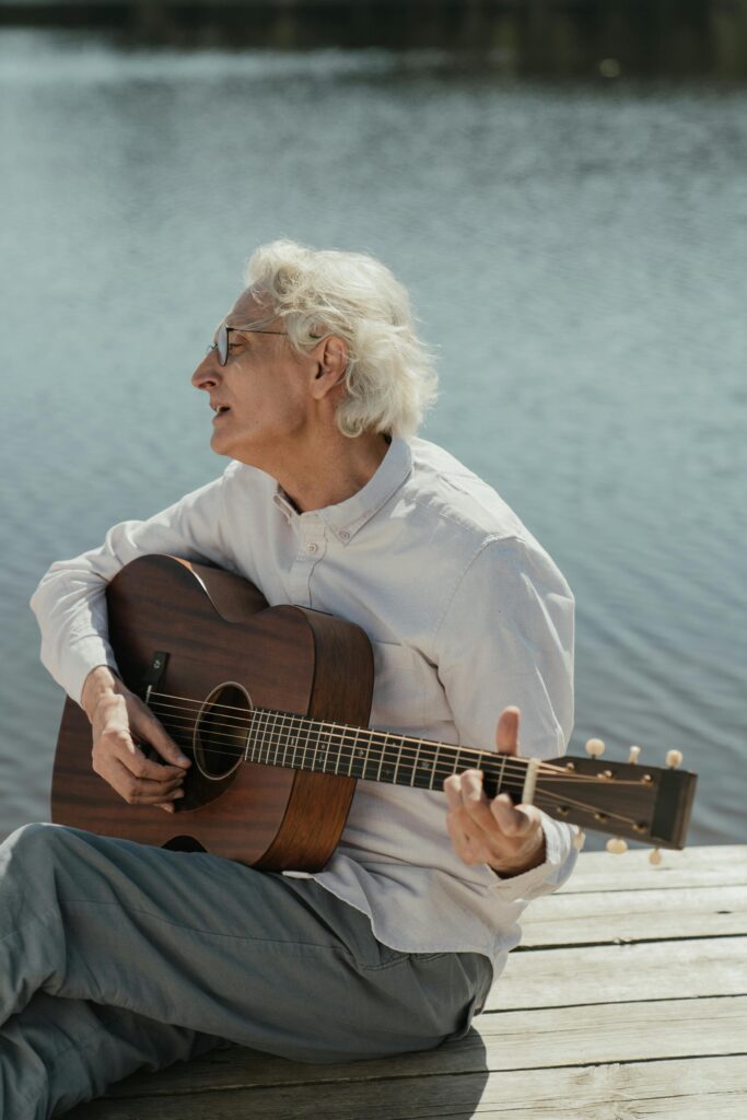 Senior man playing an acoustic guitar on a wooden dock by a calm lake, enjoying nature.