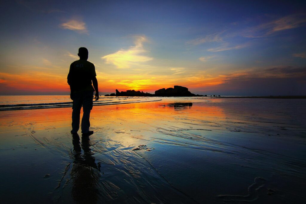 A silhouette of a person stands on the beach reflecting the vibrant sunset sky and tranquil sea.