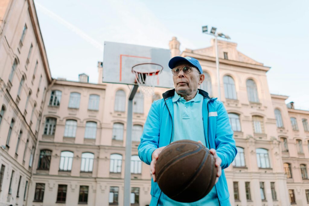 Elderly man playing basketball on an outdoor court against a city backdrop.