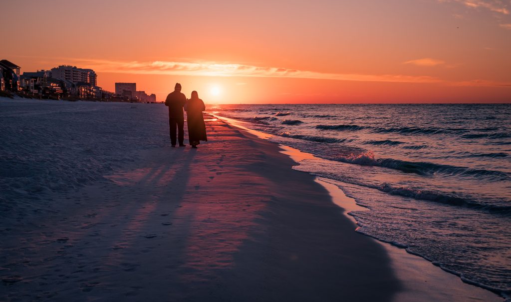 Two people walking along the beach at sunset, silhouetted against the colorful sky.