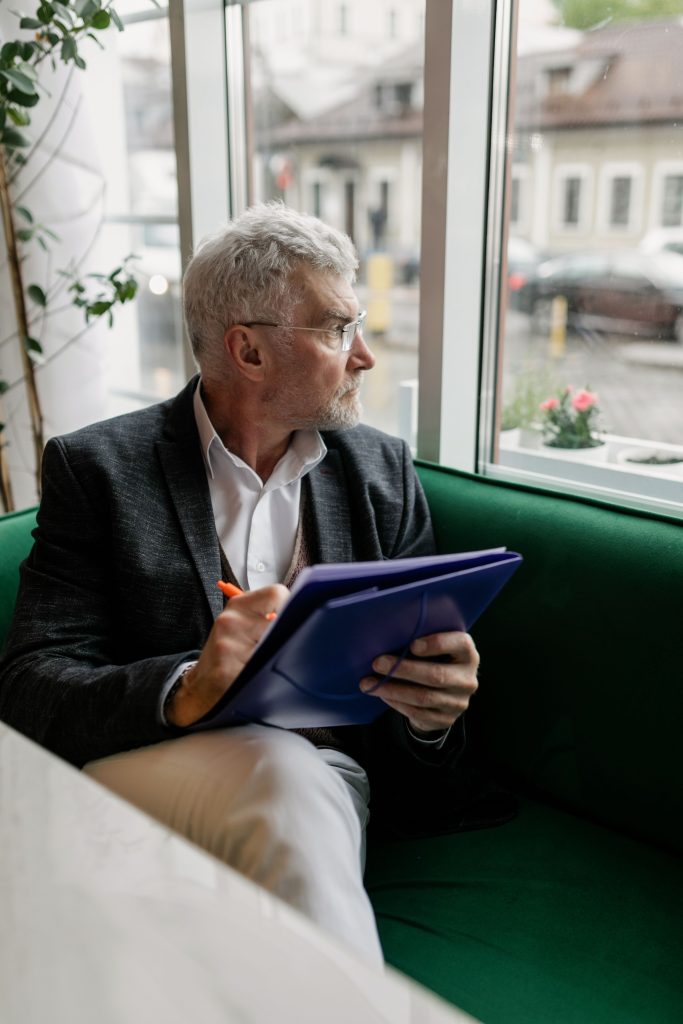 Senior man in a suit writing in a notebook while seated by café window.