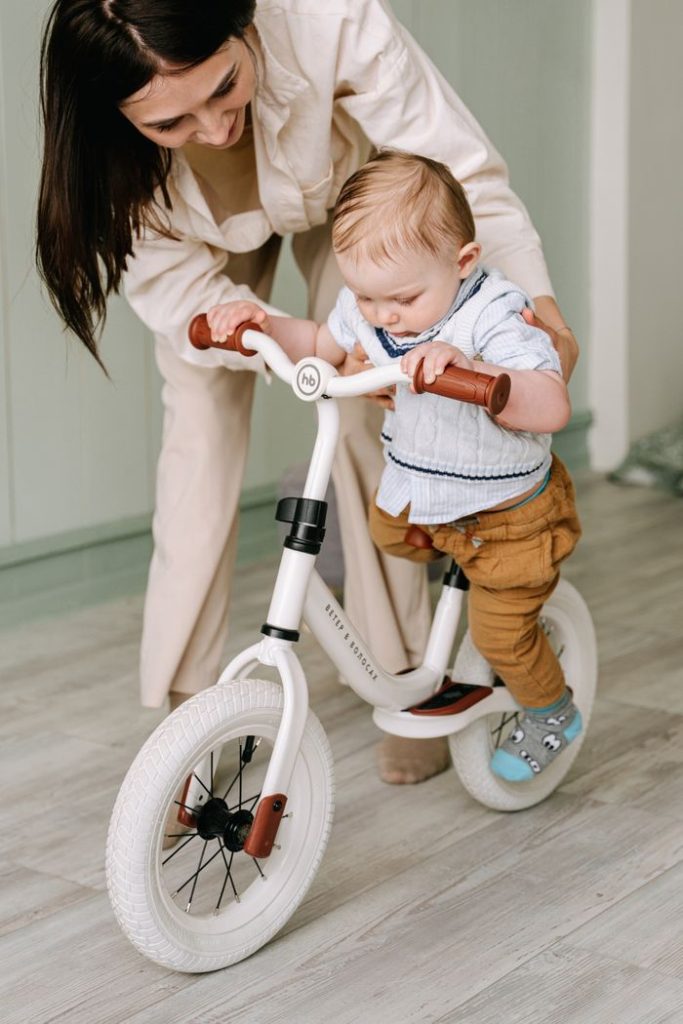 A joyful moment of a mother teaching her toddler to ride a balance bike indoors.